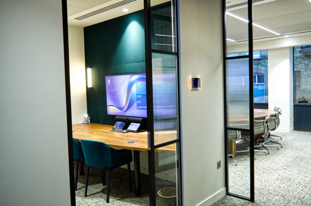Contemporary glazed meeting room with wooden table, teal chairs, wall-mounted display screen, and tabletop touch control device for video conferencing.